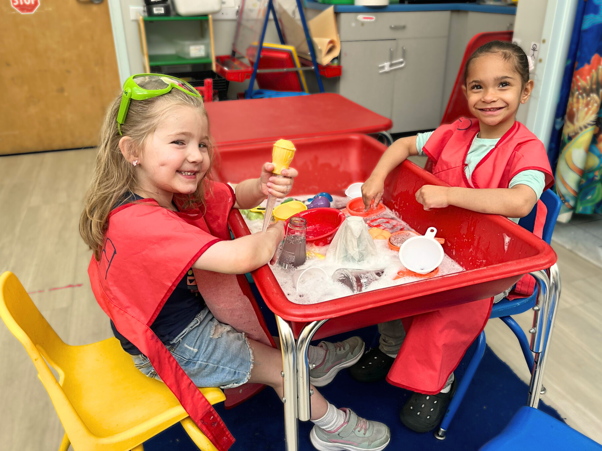 Two preschool girls in red aprons sit in chairs across from each other playing in a red water table smiling during full-day child care program