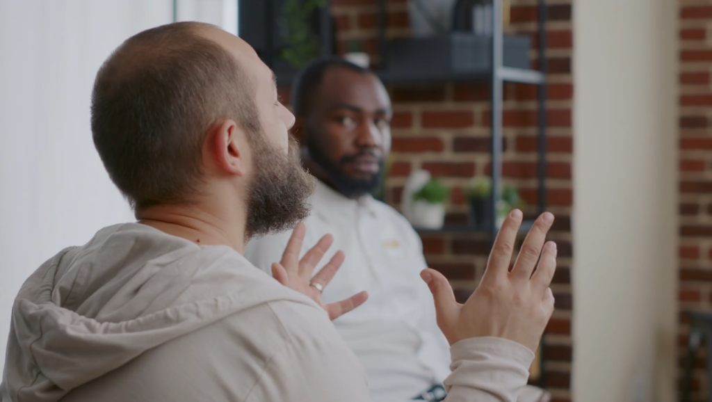 A man talks animatedly with his hands, while another man sits next to him listening and nodding. 

Caring Gift of Healing