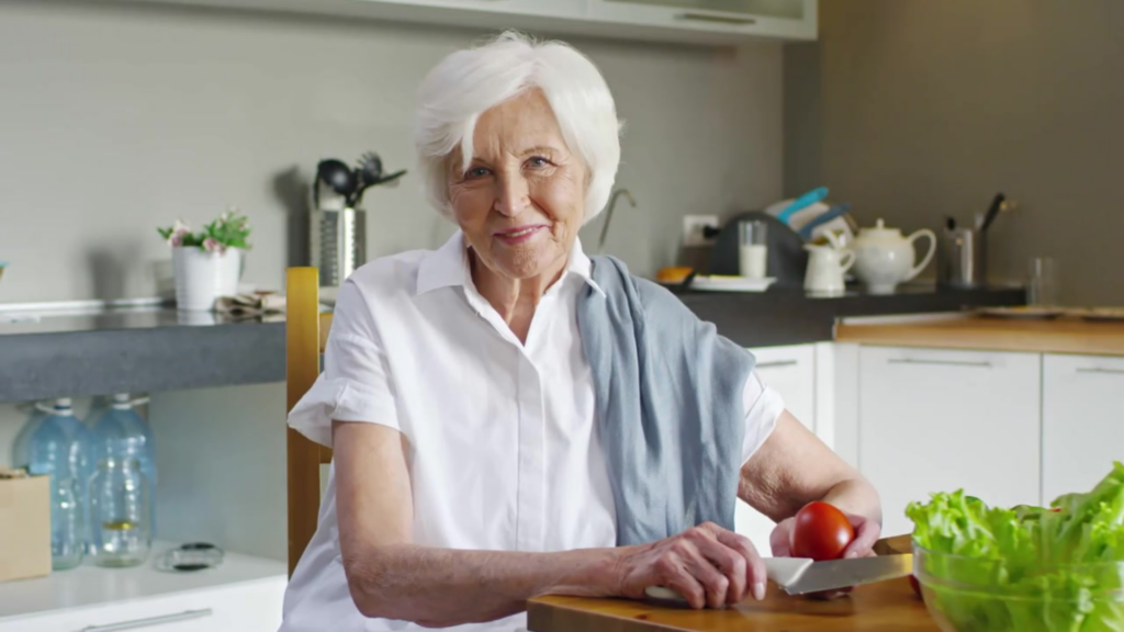 Older woman with white hair and a white shirt sits at a table in her own kitchen preparing a salad.

Caring Gift of Independence